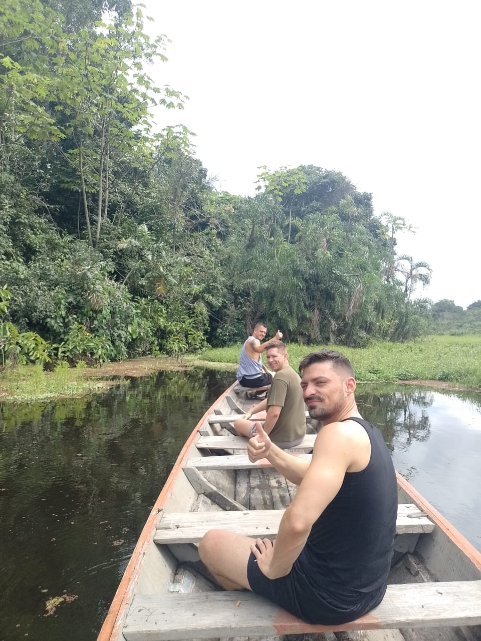 Tres turistas masculinos disfrutando de una navegación en canoa tradicional por un afluente de aguas tranquilas rodeado de densa vegetación amazónica.