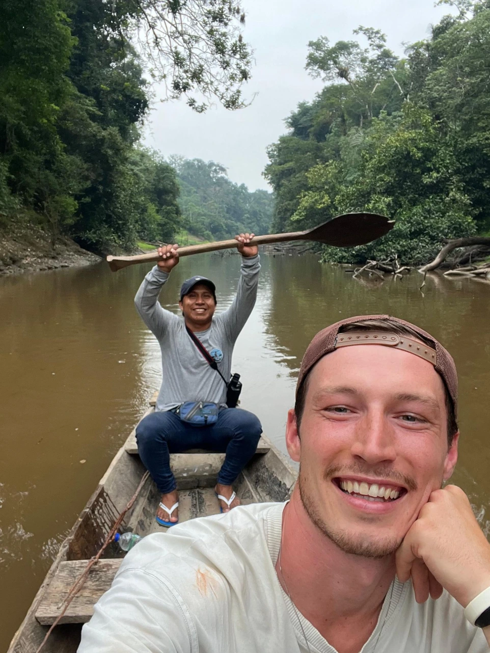 Un explorador sonriente junto a su guía local, quien sostiene un remo artesanal en alto desde una canoa tradicional, navegando por un río tranquilo durante una expedición matutina en el Amazonas.