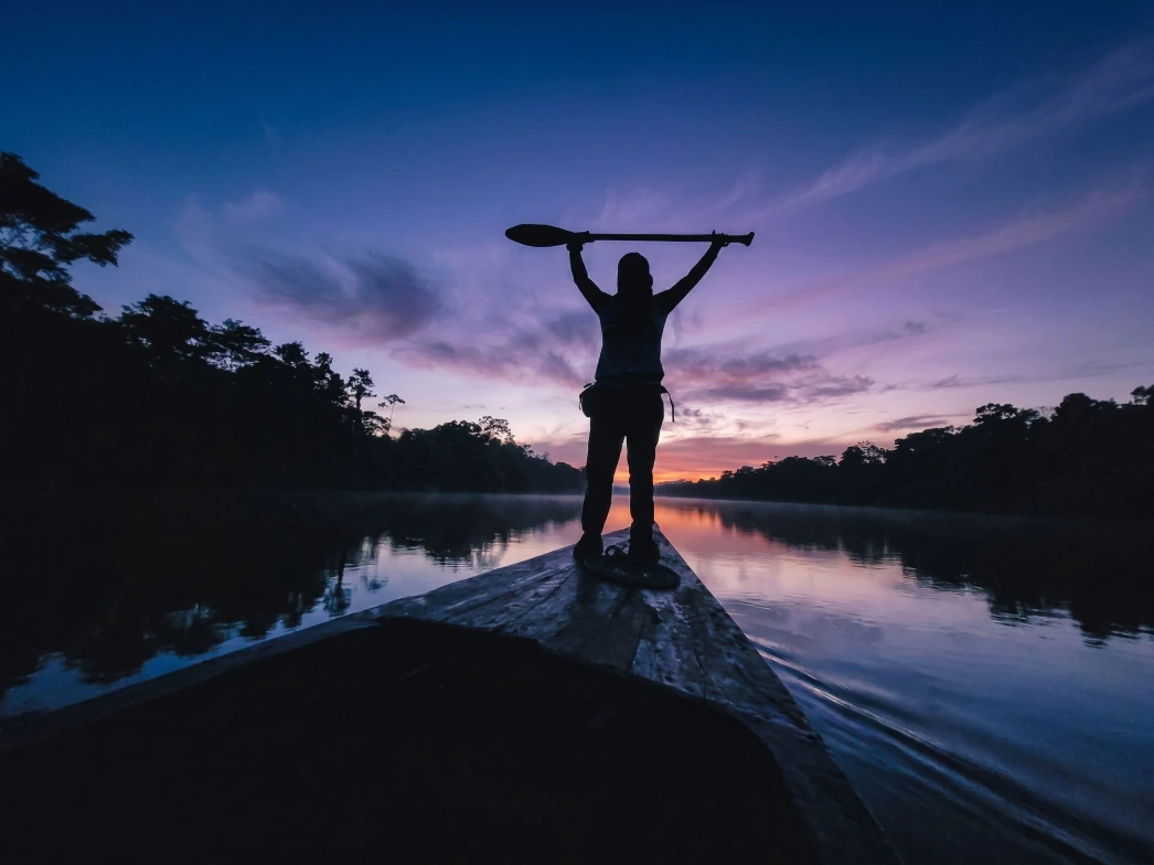 Amazon Expedition in Iquitos, Peru: a traveler standing on a traditional Amazonian boat at sunset, holding a paddle as the boat navigates calm rainforest waters. The scene captures the spirit of adventure, river exploration, and immersion in the landscapes of the Peruvian Amazon.